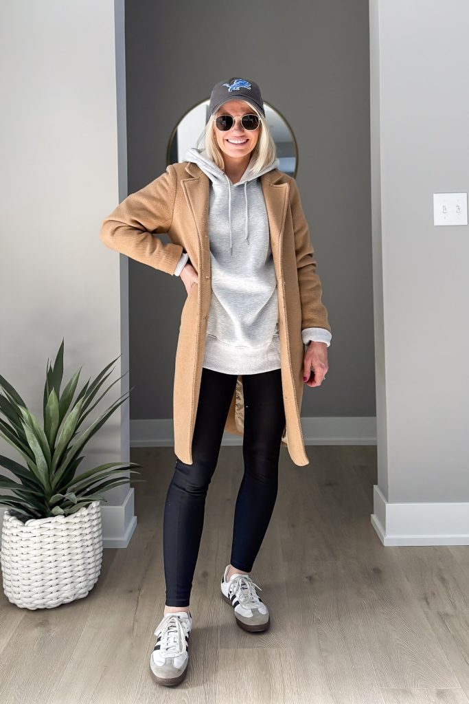 Woman in a camel coat, gray hoodie, black leggings, and sneakers, accessorized with a baseball cap and sunglasses.