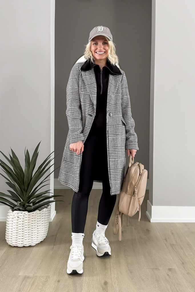 Woman in a houndstooth coat, black outfit, and white sneakers, holding a beige backpack and wearing a baseball cap.